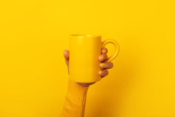 Hand with yellow nails and sweater holding a plain yellow ceramic mug in front of a solid yellow...
