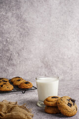 A glass of milk served with stacked chocolate chip cookies and more cookies on a cooling rack,...