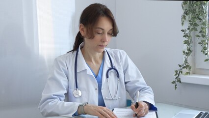 Female doctor in lab coat and stethoscope diligently writing notes on clipboard while sitting at...