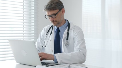 Focused mature male doctor wearing eyeglasses and lab coat with stethoscope is typing on laptop...