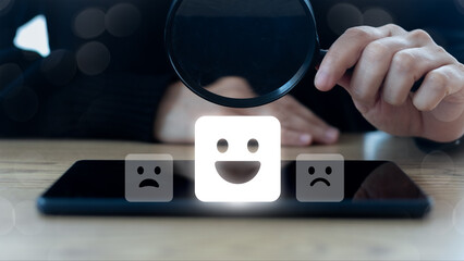 Close-up of a hand holding a magnifying glass over a happy face icon on a smartphone, representing...