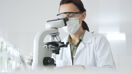 Female scientist wearing a lab coat, face mask, and safety glasses carefully adjusts a microscope,...