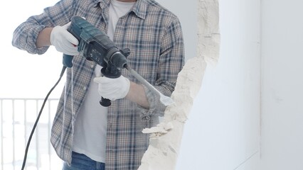 Male construction worker demolishing white wall with rotary hammer drill is wearing grey checkered...