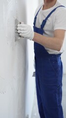 Unknown man construction worker wearing gloves and blue overalls is using a putty knife to apply...