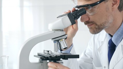 Adult male scientific researcher analyzing sample through microscope, wearing protective gear in high tech laboratory environment. Healthcare, medicine and science concept