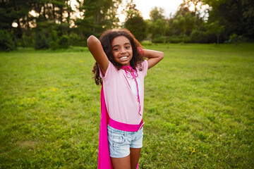 Cheerful young girl dressed in a playful pink cape standing outdoors in a bright green park,...