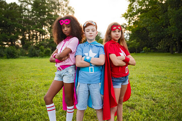 Three children wearing superhero costumes stand confidently in a park setting, embracing fun and...