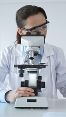 Female scientist wearing lab coat and protective glasses using microscope, analyzing samples in...