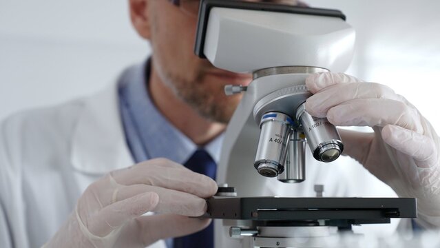 Close up of scientific researcher wearing white protective gloves carefully calibrating laboratory microscope, performing precision medical investigation. Medicine and science concept