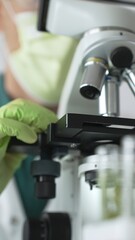 Close up of a scientist in green gloves and a mask using a microscope for research and analysis in...