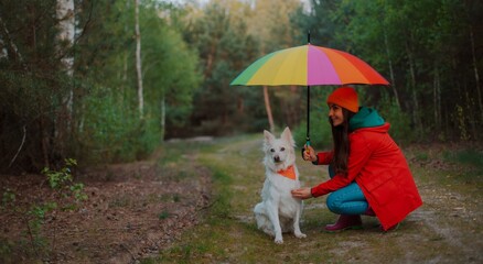 A young woman in a red coat and orange beanie is crouching next to her white dog wearing an orange...