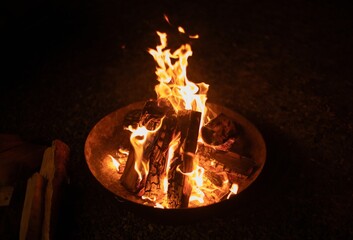 A close-up of a bright campfire burning in a metal fire pit at night. Flames dance around stacked logs, creating a warm and inviting atmosphere for camping or outdoor gatherings.