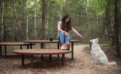Smiling young woman sits on a round wooden bench in a forest, playfully reaching out with a stick...