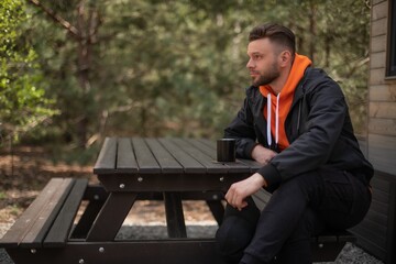 Young man in a black jacket and orange hoodie sits at a wooden picnic table outdoors, holding a cup and gazing thoughtfully into the distance in a forest setting.