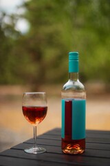 A bottle of rosé wine with a turquoise label and a filled wine glass on a dark wooden table outdoors with a blurred green background. Ideal for leisure, celebration, and outdoor dining themes.