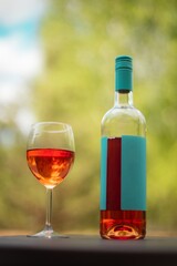 A glass of rosé wine and a matching bottle with a turquoise label set outdoors against a blurred green background. Perfect for themes of relaxation, celebration, and outdoor dining.