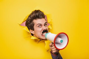 Person yelling into a megaphone while emerging through a yellow paper wall, symbolizing communication, attention-grabbing, and expression of boldness and creativity. Vibrant and attention-focused