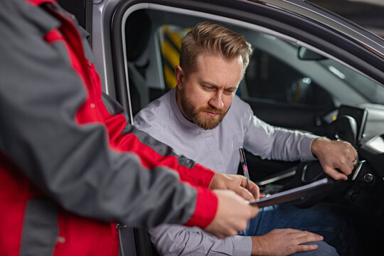 A man seated inside a car reviews and signs documents provided by an automotive professional wearing red overalls in a workshop.