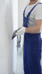Unknown man construction worker wearing gloves and blue overalls is using a putty knife to apply...