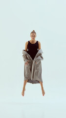 Contemporary dancer floating in mid-air with focused gaze and trench coat against light backdrop....