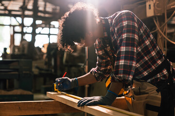Young African American Carpenter Measuring Wood Plank with Tape and Pencil, Working in Sawmill...