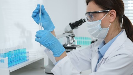 Female scientist wearing a face mask and blue gloves, carefully dropping liquid from a pipette into...