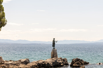 Maiden standing on a rock by the sea, holding a seagull, with a serene ocean backdrop and distant...