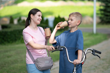 Obraz premium Mother and son walking with a bicycle in a park. The boy is drinking from a paper cup while the mother smiles and holds the handlebars