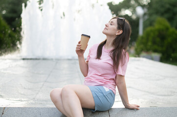 Young woman enjoying a coffee by the fountain, eyes closed, relaxed and smiling. Wearing a pink...