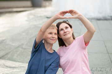 Mother and son making a heart shape with their hands while smiling and looking up. Sweet moment of...