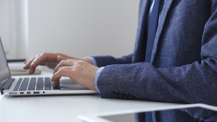 Professional businessman typing on laptop keyboard, working intently while wearing tailored blue suit and wristwatch in contemporary workspace background. Business people concept