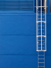 Blue walls with grooves and louver with a white steel ladder and round safety fence of an entrance to mechanical storage room for maintenance and repairs.