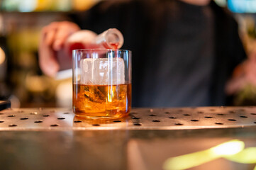 Close-up of bartender pouring golden liquor into glass with ice cubes on metal bar counter, showcasing drink preparation in stylish nightlife setting.