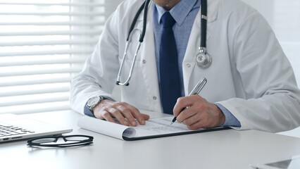 Close-up of male doctor's hands filling out medical form, sitting at desk with laptop and glasses, wearing white coat and stethoscope in medical office. Medicine and healthcare concept