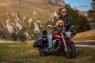 Happy biker couple posing with red cruiser motorcycle in scenic mountain setting during golden...