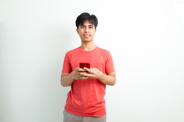 Smiling young Asian man in orange t-shirt using smartphone on white isolated background, perfect...