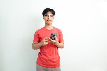 Smiling young Asian man in orange t-shirt using smartphone on white isolated background, perfect...