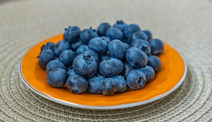 Fresh ripe blueberries in a bright orange bowl on a textured woven placemat. Top view of a healthy superfood snack, rich in vitamins and antioxidants. Concept of healthy eating, summer fruit harvest