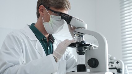 Adult male scientist wearing white mask, lab coat and protective glasses using microscope, doing...