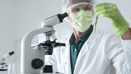 Adult male scientist wearing green protective gear looking at plant samples in test tubes and using...