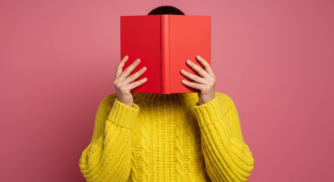 A person wearing a yellow sweater hides their face behind a red book