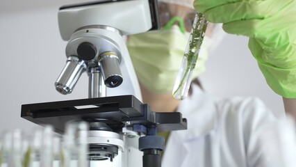 Female scientist wearing green protective mask and gloves is holding test tube with plant sample,...