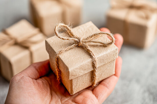 Hand holding a small wrapped gift box with a rustic twine bow, surrounded by similar packages on a neutral background