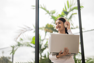 Young asian businesswoman listening music with headphones and holding laptop near window