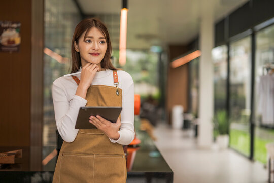 Young thoughtful asian barista is holding tablet and stylus in cafe