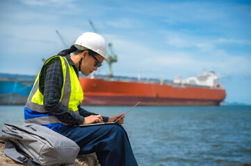 Female ship engineer sitting on shore working  tablet with marine ship in the sea