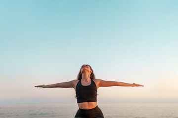 Woman wearing black sportswear practicing yoga pose on sandy beach, stretching arms overhead during...