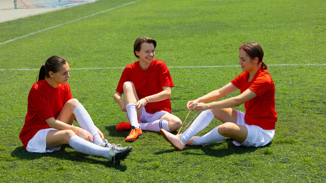 Three female soccer players seated on grass getting ready for a match. Concept of mental and physical preparation, active teamwork warmup, sports training for girls and young women. - Powered by Adobe
