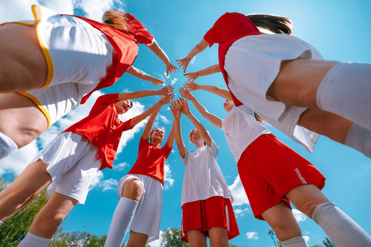 Girls football team in red and white forming team huddle with hands raised under blue sky. Concept of sports unity, youth motivation, girl empowerment, summer team building. - Powered by Adobe