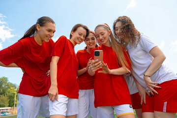 Group of young female soccer players in red and white jerseys smiling while looking at phone outdoors. Concept of youth bonding, team celebration, social sharing, summer sports lifestyle. © master1305
